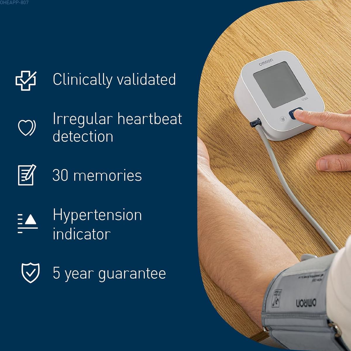 White OMRON M2 blood pressure monitor on a wooden table with a user wearing the grey upper arm cuff and pressing the button.