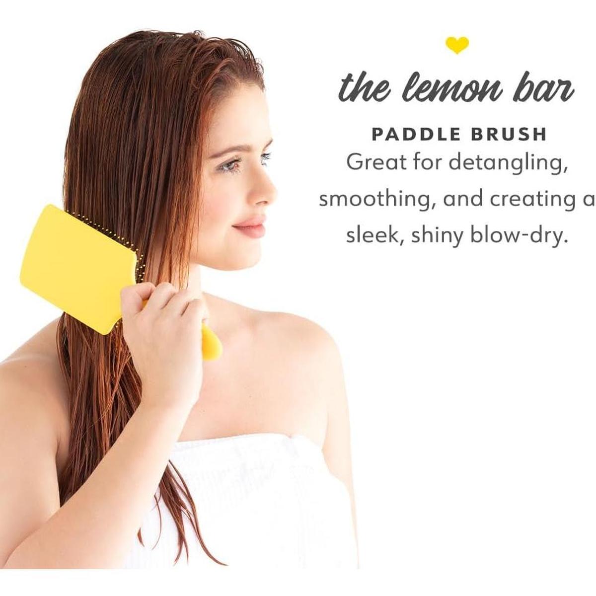 Woman in a white towel using a bright yellow Drybar paddle brush to detangle her long, damp hair.