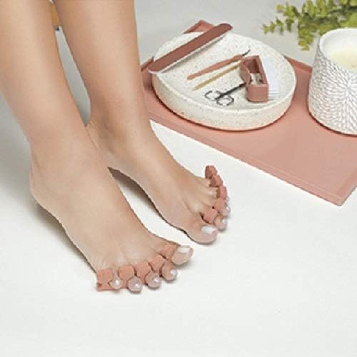 Feet wearing pink toe separators next to a Revlon pedicure kit with nail tools on a pink tray.
