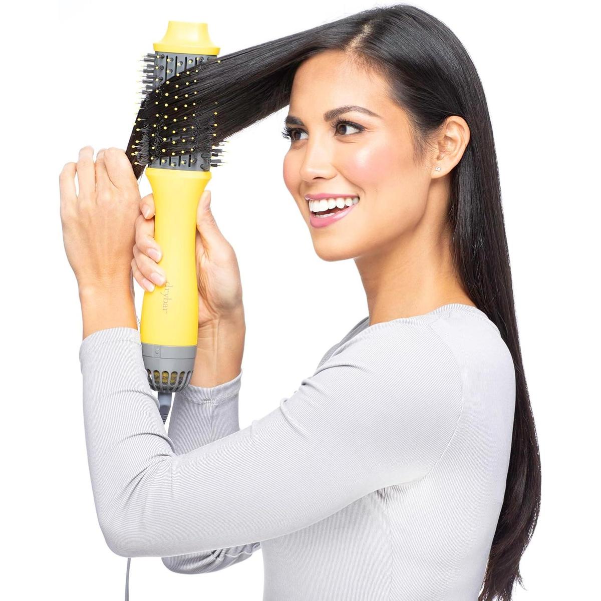Woman styling long dark hair with a yellow Drybar Double Shot 6.1cm oval hot air brush with tufted bristles.